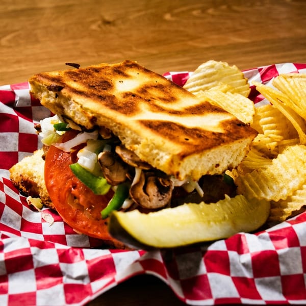 Grilled sandwich with vegetables and mushrooms, served with crinkle-cut chips and a pickle.
